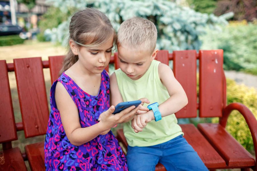 Happy kids sitting on bench and using smartwatches with interest, data synchronization between smartphone and smartwatch, new technology for children, digital education and care, outdoors lifestyle